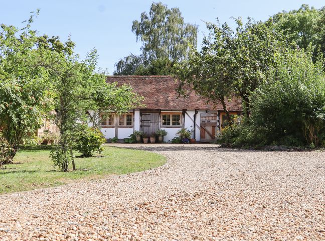 A gravel driveway leading to a small house with wooden doors and windows surrounded by trees and shrubs at The Old Stables in Headcorn
