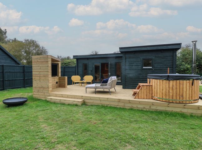 A wooden deck with outdoor seating a hot tub and a barbecue under a partly cloudy sky at Badger's Burrow in South Luffenham near Edith Weston