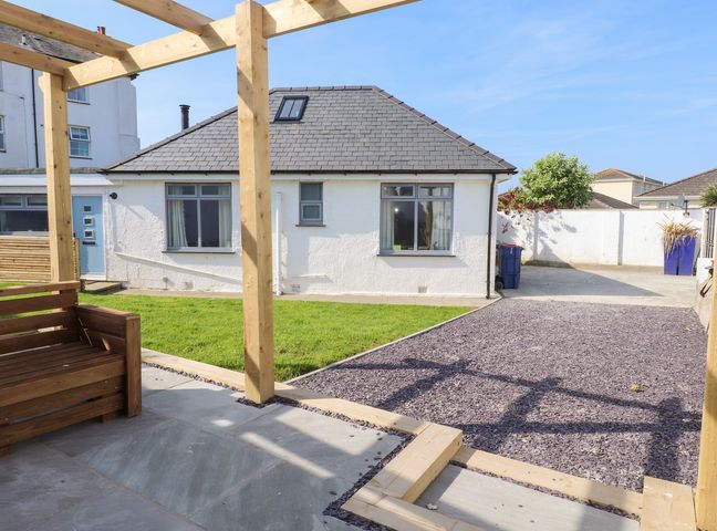 A backyard with a wooden pergola and seating area a gravel path a green lawn and a white house at Trearddur House Cottage in Trearddur Bay
