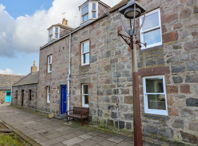 A stone building with white windows and a blue door beside a wooden bench and a street lamp at Footdee Cottage Aberdeen