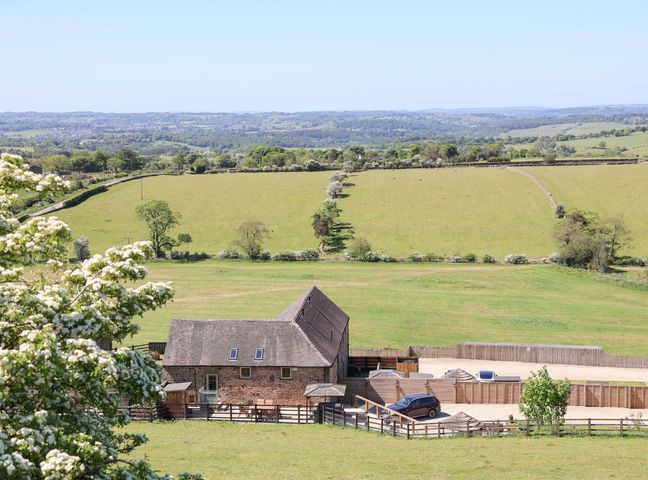 A barn and a car parked beside a fenced area with green fields and trees in the background at Parlour Barn in Leek