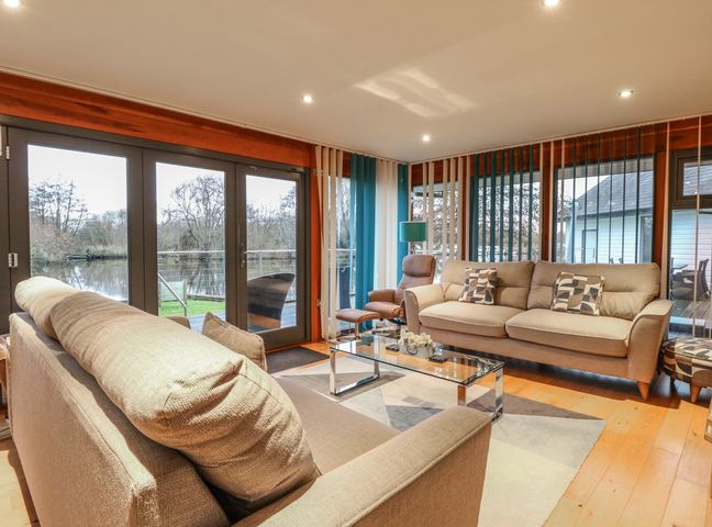 A living room with beige sofas a glass coffee table and large windows overlooking water at Tealby in Horning