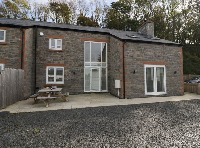 The exterior of a stone house with white framed windows and doors with a wooden picnic table on a patio at Edgemere Cottage in Cartmel