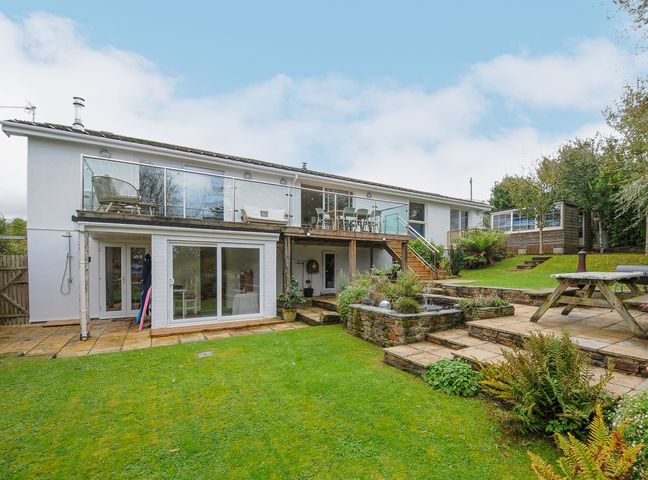 A backyard with a grassy lawn a wooden deck with glass railing patio furniture and a stone planter at Seamay in Polzeath