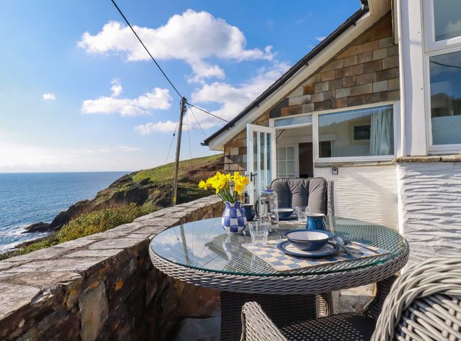 An outdoor stone terrace with a round glass table set for a meal overlooking a coastal view at Suntrap House in Portloe