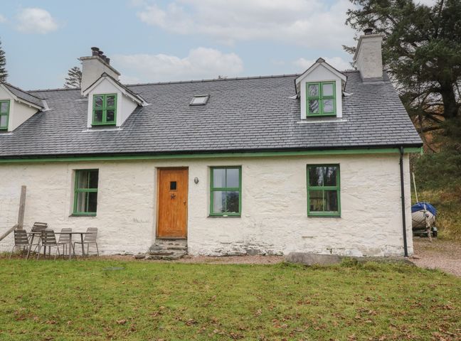 A white cottage with green window frames and a wooden door with outdoor chairs and table on grass at Woodland Cottage in Druimindarroch near Arisaig