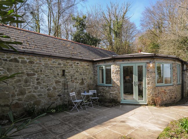An outdoor stone patio with two white chairs and a small table in front of a stone building with blue framed windows and doors at Rose Cottage in Callington
