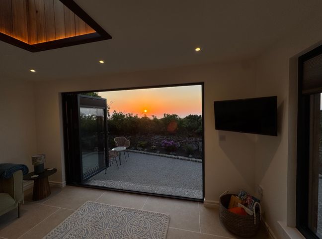 A room with a rug on the floor and a wall-mounted TV next to an open large door showing an outdoor patio at sunset at Little Victory in Pendeen