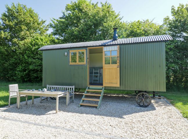 A green corrugated metal shepherd hut with open door and window on a gravel patio with wooden table chairs and bench surrounded by trees at Hazel South Leigh near Witney