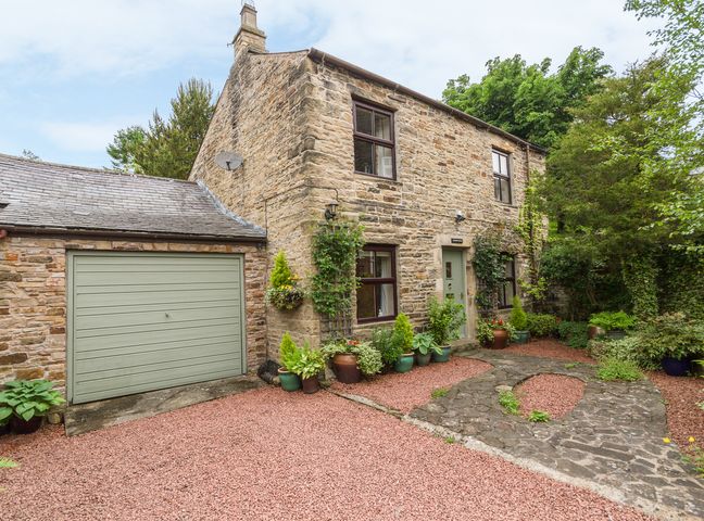 A stone cottage with a green garage door and potted plants outside surrounded by trees at Crescent Cottage in Haltwhistle near Hadrian's Wall