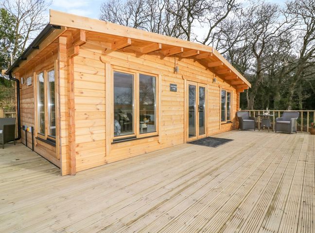 A wooden cabin with large glass windows and doors on a deck with outdoor chairs and a table at Little Orchard Lodge in Goonhavern