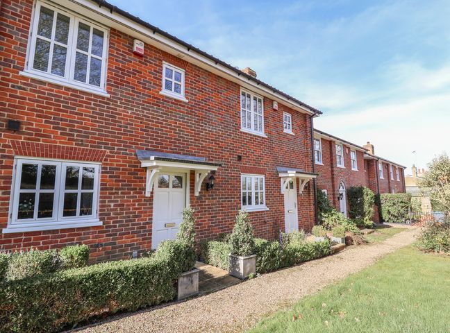 A row of red brick townhouses with white doors and windows along a gravel path with trimmed hedges and grass