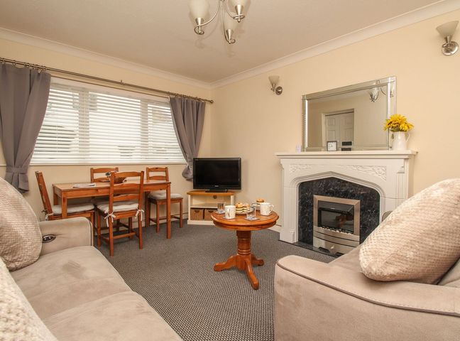 A living room with a small wooden dining table and chairs a round wooden coffee table a fireplace with a mirror above a television on a stand and beige sofas at Seagulls Nest in West Bay
