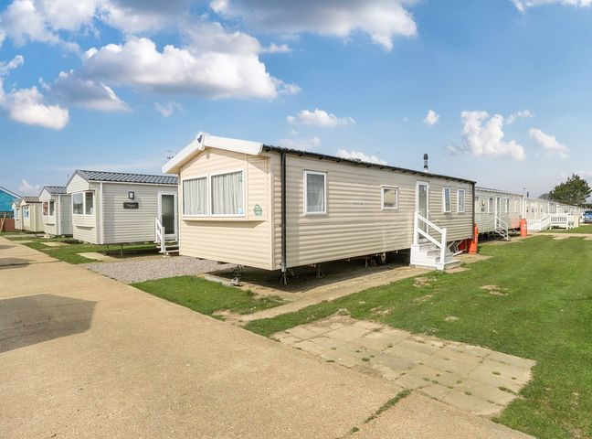 A row of beige and white mobile homes on grassy plots with a paved path and blue sky at Ripples & Reeds - Billing Aquadrome in Billing Aquadrome Holiday Park