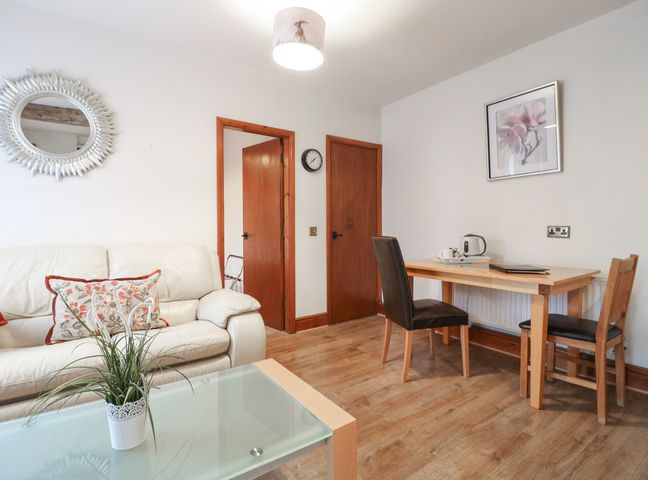 A living area with a white sofa and floral cushion a glass coffee table with a plant a wooden dining table with two chairs a wall clock and framed artwork at Stables in Castle Donington