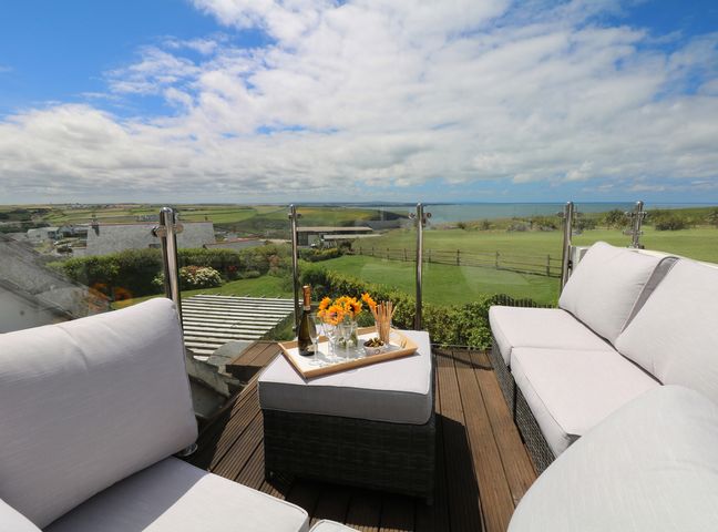 An outdoor seating area with white cushioned chairs and a table with drinks and flowers overlooking fields and the sea at Coastmans Nest in Mawgan Porth