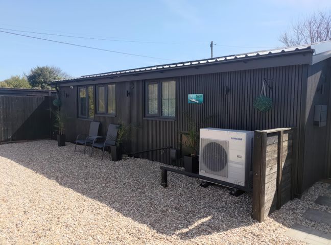 A dark exterior wall with two chairs and potted plants in front and an air conditioning unit at 19 The Court in Anderby Creek