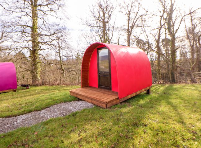A small red pod with a wooden door and a wooden deck on grass with leafless trees in the background at Pod 4 in St Asaph