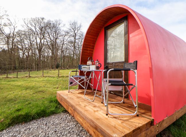 A small curved red cabin with a wooden door and two metal chairs with a small table on a wooden platform outside in a grassy area at Pod 5 in St Asaph