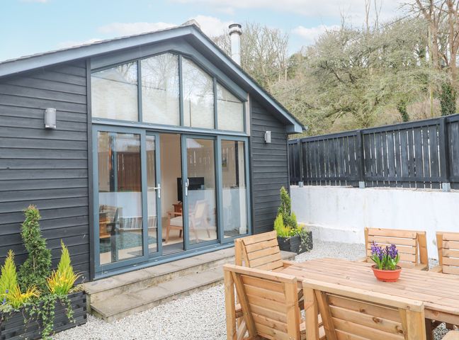 A patio with wooden chairs and table with potted plant outside a black wooden building with large glass doors at Chy Benyngoth in Hayle