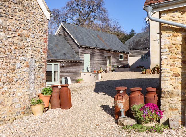 A courtyard with gravel ground surrounded by stone and wooden buildings with old milk churns and potted plants at The Dairy at Wyke Farm in Wyke near Lyme Regis