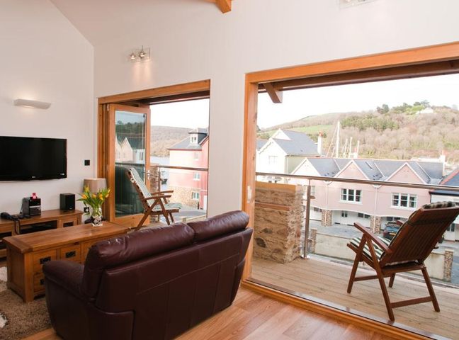 A living room with a brown leather sofa wooden furniture a wall-mounted TV and chairs on a balcony overlooking houses and hills at Quay Lodge in Dartmouth