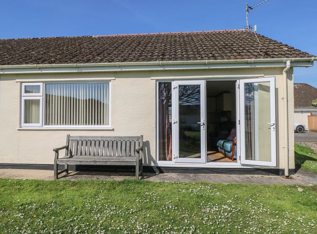 The exterior of a single-story house with a wooden bench on a concrete path and open double glass doors showing a blue armchair inside at Kites - 52 Gower Holiday Village in Scurlage near Port Eynon and Rhossili