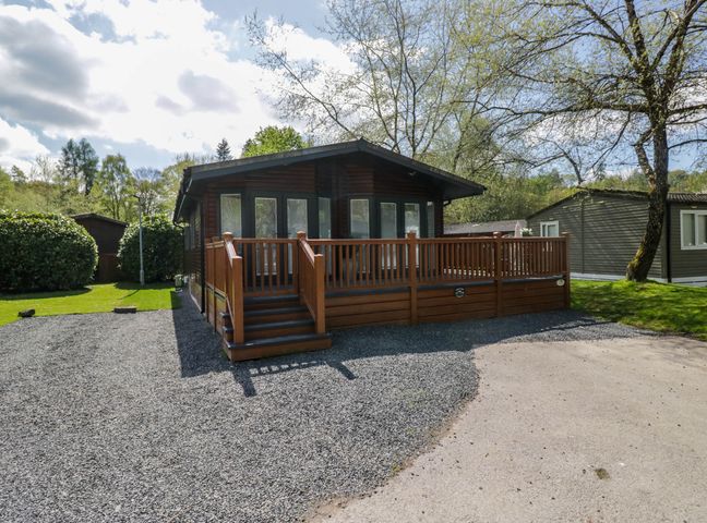 A wooden lodge with a deck and stairs surrounded by gravel and grass with trees nearby at Robins Lodge in White Cross Bay near Troutbeck Bridge