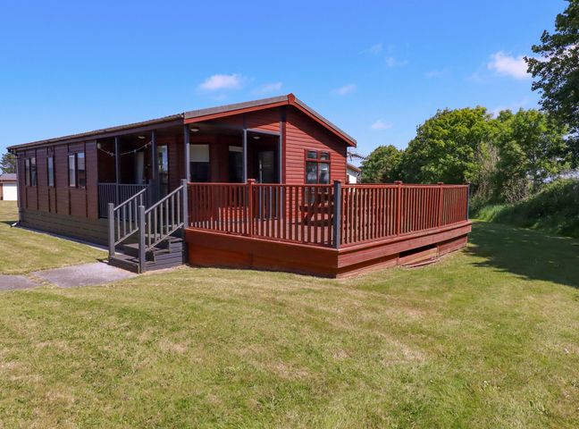 A wooden cabin with a fenced deck and steps on a lawn with trees in the background at Lodge 9- Lamorna in Tregolds near St Merryn