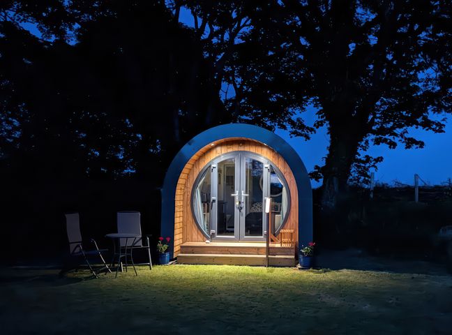A small round wooden pod with glass double doors lit at night with two chairs and a table outside at Florence Pod in Pembroke