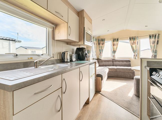 A kitchen area with cupboards sink kettle toaster and microwave leading to a living area with sofa windows and curtains at Golden Dune - Hayling Island in Hayling Island Holiday Park