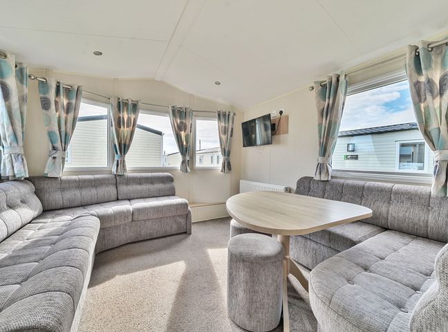 A living room with grey upholstered seating a wooden table and multiple windows with patterned curtains at The Lookout - Hayling Island in Hayling Island Holiday Park