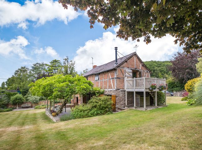 A brick and stone house with a wooden balcony surrounded by grass and trees at The Cider Mill in Westhope near Leominster