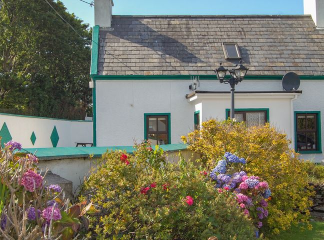 A white house with green trim and a slate roof with flowers and shrubs in the garden at Nana's House in Allihies County Cork