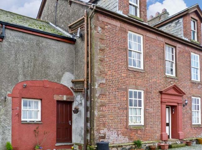 The exterior of a brick house with multiple windows and two doors one door has a red frame the other has a red border and both have small potted plants nearby at Gable View in Ravenglass