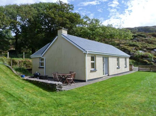 A single-story house with a stone patio and wooden chairs and a table on a grassy lawn at The Bay in Caherdaniel County Kerry