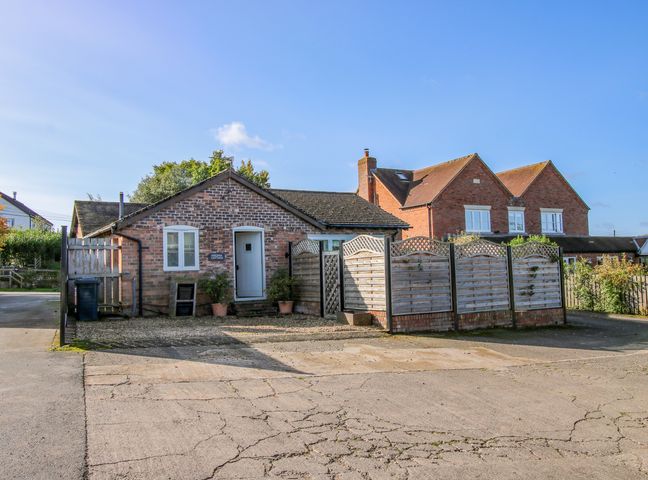 A small brick cottage with a white door and windows next to a wooden fence and a larger brick house in the background at Virginia Cottage in Acton Burnell near Much Wenlock