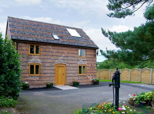 A two-story brick house with wooden framed windows and door a paved driveway with a black water pump surrounded by flowers and a wooden fence at The Old Cider Mill in Caynham near Ludlow