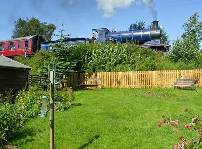 A red and blue steam train passing behind a garden with a wooden fence and benches at Steam Cottage in Aviemore