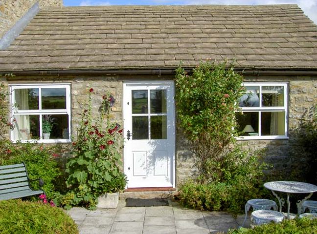 A stone cottage with a white door and two windows with plants and flowers outside at Curlew Cottage in Barnard Castle