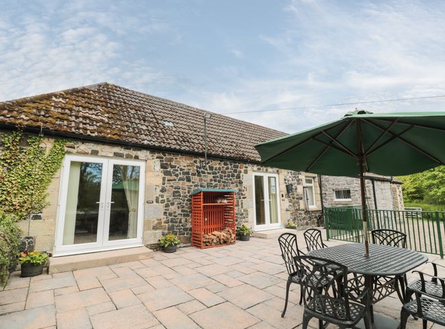 A stone patio with black metal table and chairs under a green umbrella at Gardener's Cottage in Belford near Bamburgh