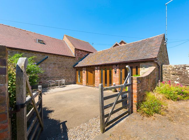 An open wooden gate leading to a paved courtyard with two metal chairs and a table beside a stone and brick building at The Byre in Billingsley near Bridgnorth