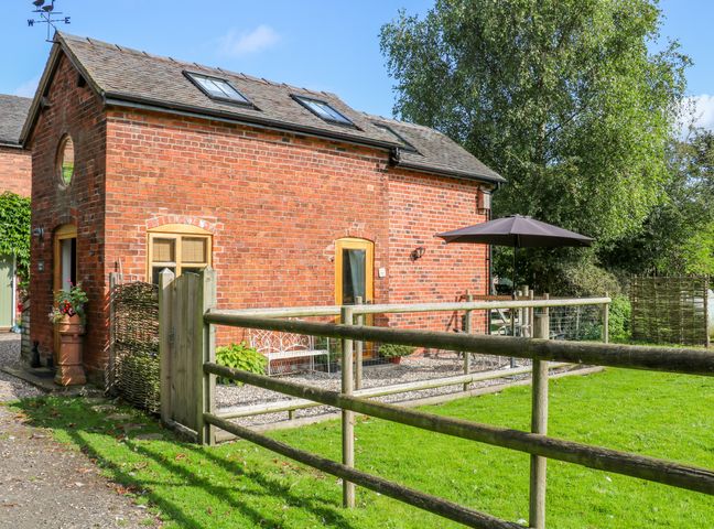 A small red brick building with wooden framed windows and skylights a wooden fence and gate grass and trees at Chequer Stable in Sandbach