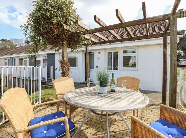 An outdoor patio with a round wooden table and four wicker chairs with blue cushions under a wooden pergola at 8 Munday Cottages in Yarmouth