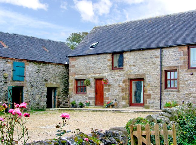 A stone building with wooden doors and windows and pink flowers in front at Aunt Janes in Winster