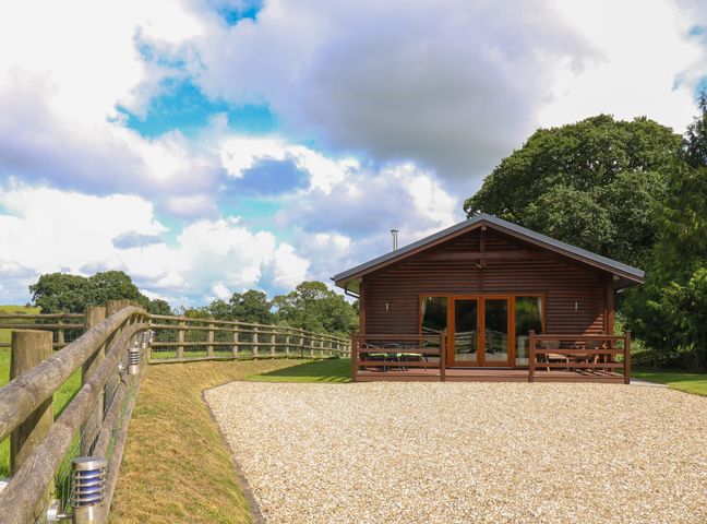 A wooden lodge with a deck surrounded by a gravel area and wooden fence in a rural setting at Barn Shelley Lodge in Copplestone near Crediton