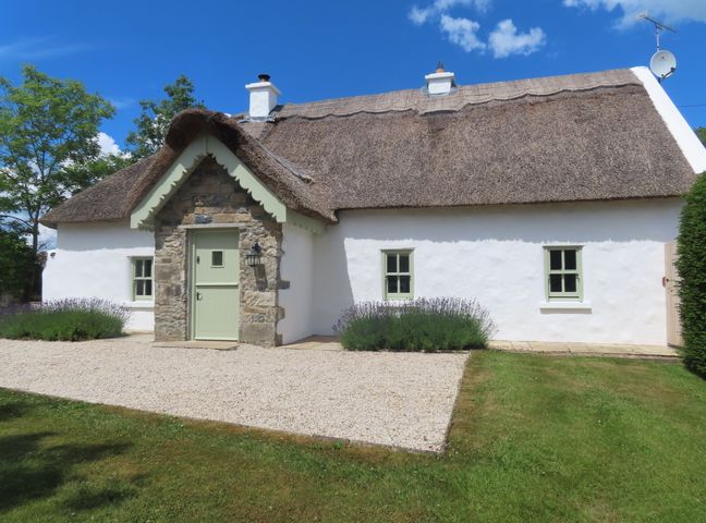 A white cottage with a thatched roof green door stone porch and small windows surrounded by grass and bushes at The Humble Daisy in Woodford County Galway