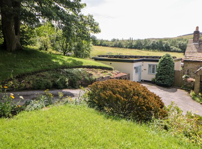 A garden with grass shrubs and flower beds next to a white building and a stone house at The Lodge in Disley