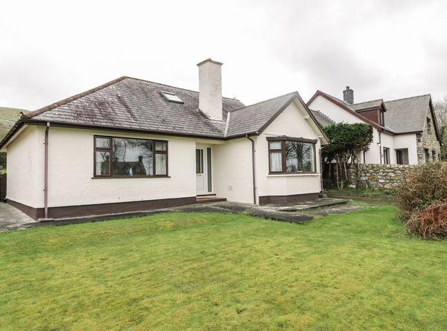 A house with a sloped roof, two large windows, a chimney, and a front door with a grass lawn at Brynteg in Llwyngwril