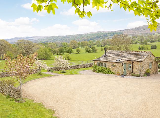 A stone cottage with a gravel driveway and trees in a rural landscape at Reynard Ing Cottage in Addingham